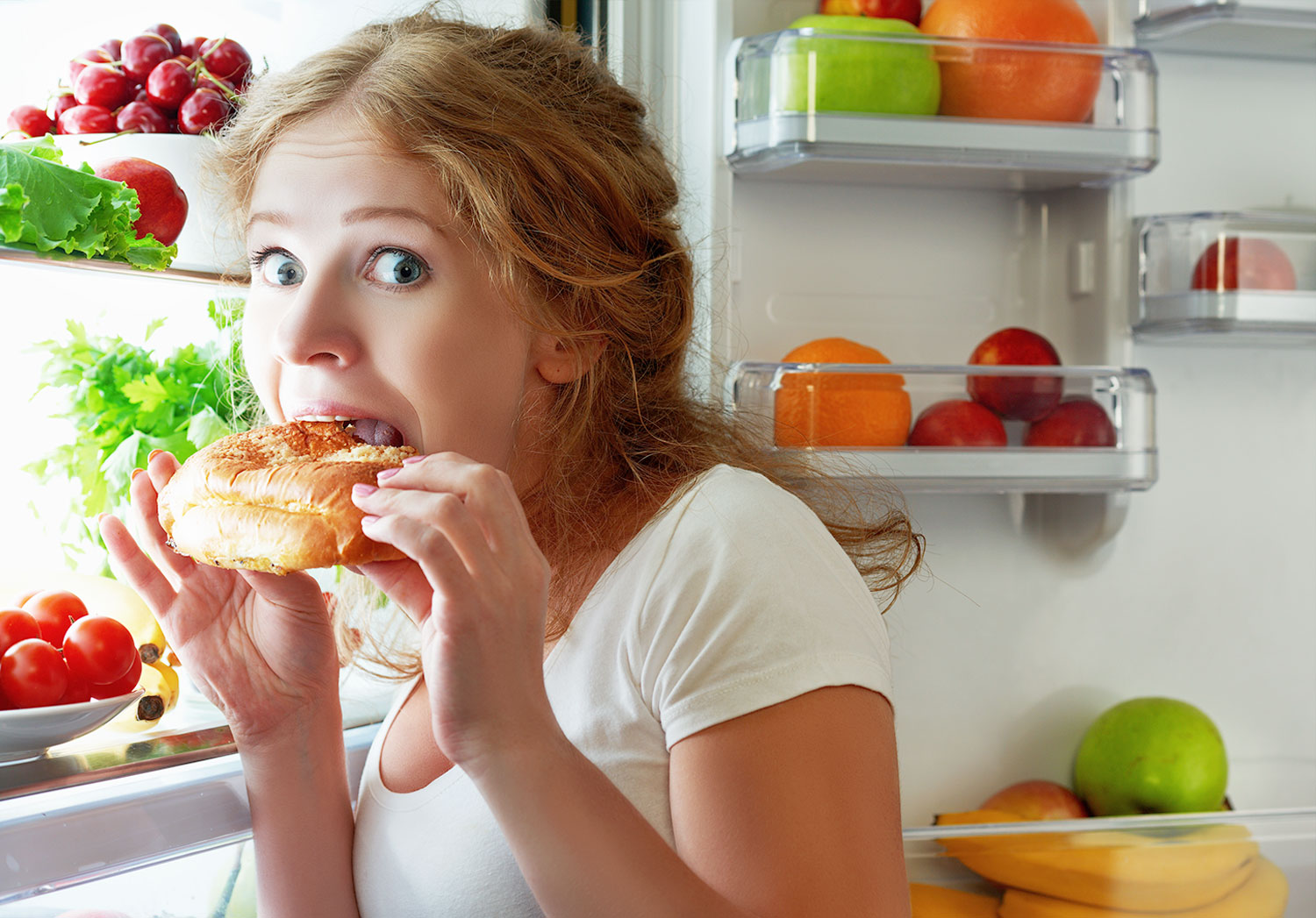 woman overeating at night caught up with burger on her hands while being in front of the fridge