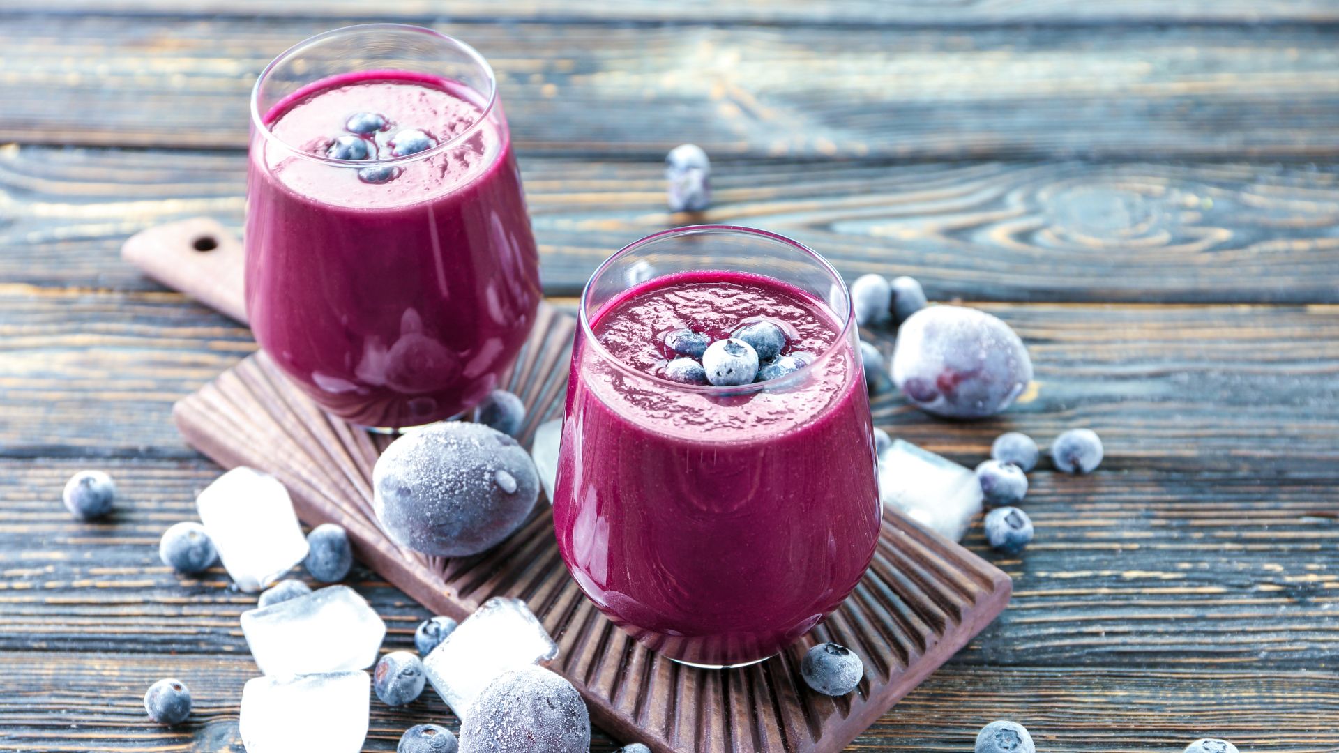 two glasses of acai juice with blue berries in them sit on top of a wooden table with frosted berries and ice cubes around them