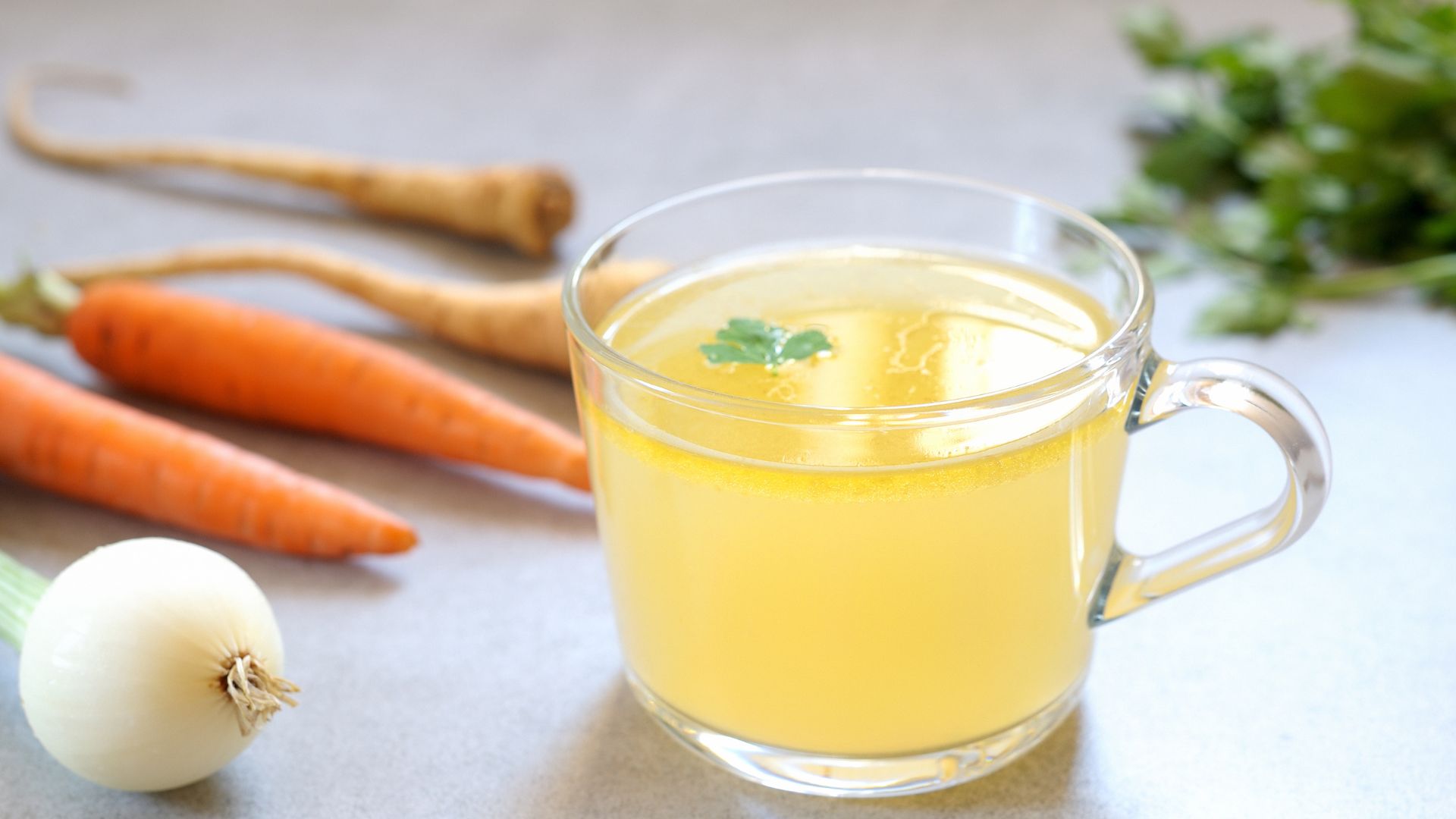 bone broth sits in a glass cup on a table with out of focus carrots and an onion in the background to help boost collagen production