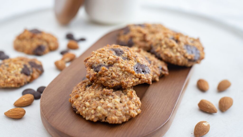 A wooden plate of healthy chocolate chip cookies with almonds scattered around.