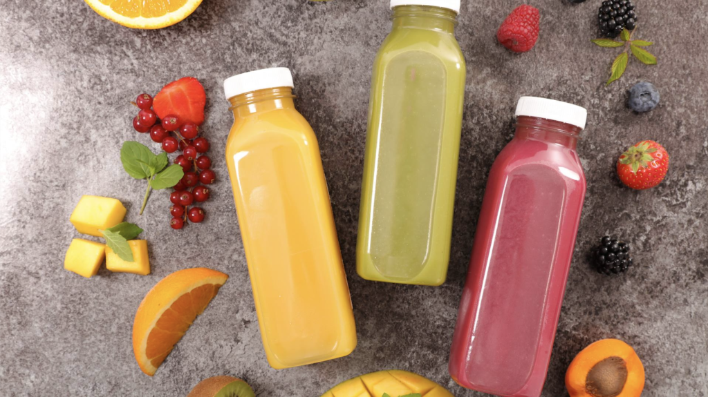 The image displays three juices in clear bottles with a white cap. They are yellow, green, and berry colored. Surrounding the juices are strawberries, blueberries, blackberries, and other fruits. The image demonstrates juicing strategies to reduce inflammation.