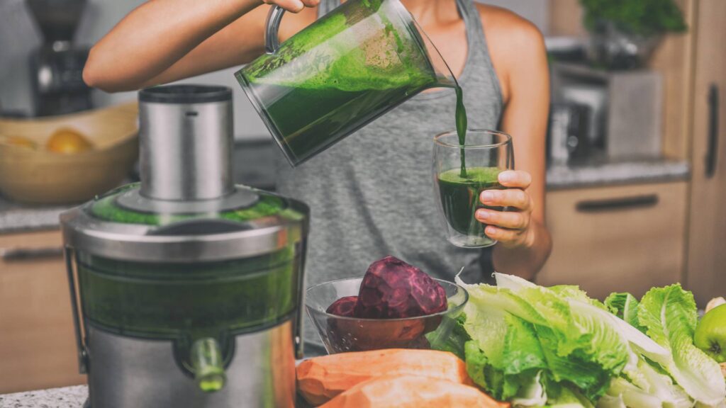 A person stands in their kitchen next to a juicer, pouring green juice into a class. They are surrounded by vegetables on the counter, demonstrating juicing recipes.